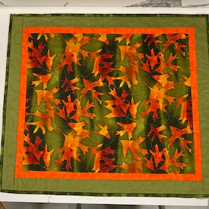 May include: A green and orange quilted table runner with a pattern of autumn leaves. The runner has a double border of green and orange fabric.