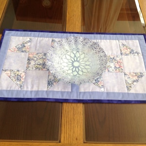 May include: A blue and white quilted table runner with a floral pattern. The runner has a blue border and is decorated with a white lace bowl in the center.