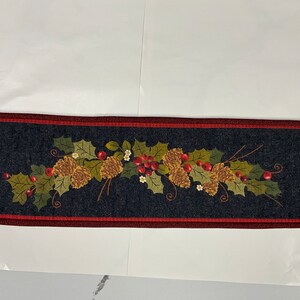 May include: A dark blue table runner with a red border features a festive design of holly leaves, pine cones, and red berries.
