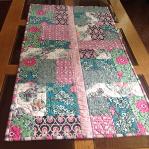 May include: A patchwork table runner with a pink, green, and blue floral pattern. The runner is made of fabric squares sewn together.