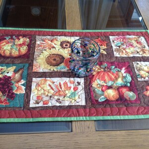 May include: A quilted table runner with a patchwork design featuring autumnal themes. The runner has a red border with a green trim and features images of pumpkins, sunflowers, leaves, and grapes. A clear glass with colorful flecks sits on top of the runner.