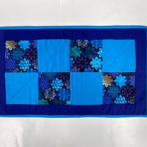 May include: A blue and purple quilted table runner with a floral pattern. The runner has a dark blue border.