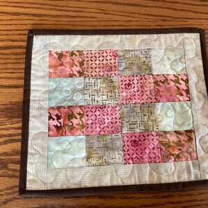 May include: A small quilted coaster with a brown border and a patchwork design of pink, brown, and white fabric squares.