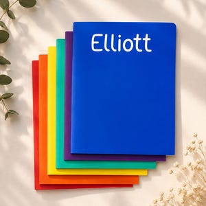 May include: A stack of six rectangular notebooks arranged in a rainbow color sequence, featuring red, orange, yellow, green, purple, and a top blue cover with the white text Elliott printed in a rounded sans-serif font on the front cover.