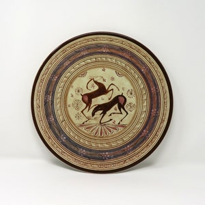 May include: A round ceramic plate with a brown and white design. The center of the plate features two stylized deer in brown and white, surrounded by a floral pattern. The outer rim of the plate has a brown and blue geometric pattern.