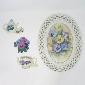 May include: A collection of floral-themed wall decor. Includes a white oval-shaped framed picture with a pansy bouquet, and three ceramic wall hangings: a teapot with cups, a cluster of pansies, and a teacup.