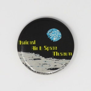 May include: A round button with a black background and a white moon with a blue earth in the sky. The text "National Air & Space Museum" is written in yellow letters on the button. The text "A Smithsonian Institution" is written in smaller letters at the bottom of the button.