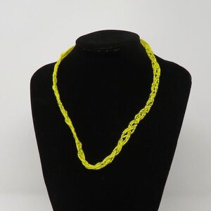 May include: A bright yellow necklace with a braided design is displayed on a black mannequin. The necklace is made of a thin, woven material and is draped around the mannequin's neck. The background is plain white.