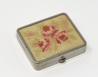Könnte beinhalten: Eine kleine, rechteckige, silberfarbene Metalldose mit einem Scharnierdeckel. Der Deckel ist mit einem floralen Muster verziert, das in rosa und braunem Garn auf einem cremefarbenen Stoff gestickt ist.
