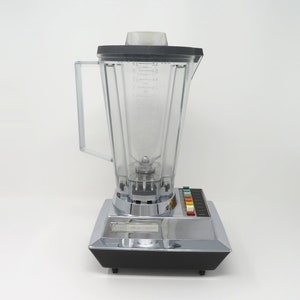 May include: A silver and black blender with a clear plastic pitcher and a control panel with multiple buttons. The blender has a silver base and a black top.