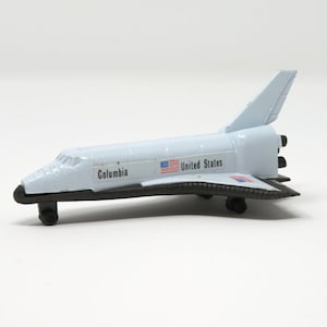 May include: A white toy space shuttle with black accents and the words "Columbia" and "United States" printed on the side. The shuttle has a small American flag printed on the side.