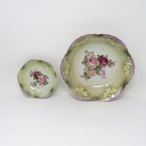 May include: A small porcelain bowl and a larger porcelain bowl, both featuring a scalloped edge with a light purple trim. The bowls have a cream-colored center with a central floral bouquet of pink and red roses, surrounded by green shading.