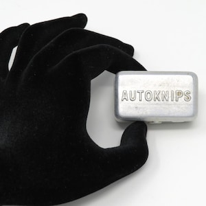 May include: A rectangular, silver-colored metal object with the word "AUTOKNIPS" embossed on its top surface. The object is held by a black glove against a white background.