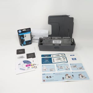 May include: An HP printer with an open top, ink cartridge, and instruction manuals. The printer is gray, and the ink cartridge is black and blue. The instruction manuals are white and blue.