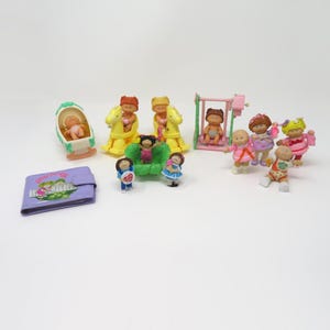 May include: A collection of miniature plastic dolls and accessories including a purple wallet with a graphic print, a cradle, two yellow rocking horses, a pink swing set, and several small figures with various hair colors and outfits.