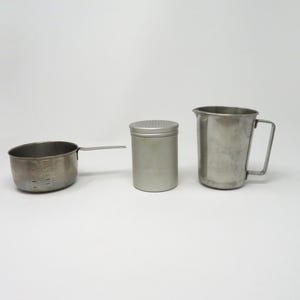 May include: A set of three kitchen tools made of stainless steel, including a small measuring cup with a long handle, a cylindrical shaker with a perforated lid, and a tall pitcher with a side handle, all featuring a metallic silver finish.