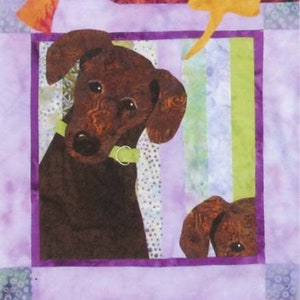 May include: A brown and black dog with a green collar is looking to the right. The dog is in a quilt square with a purple border and a multi-colored background.