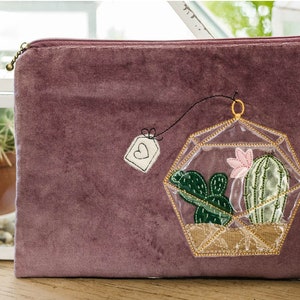 May include: A purple velvet pouch with a gold zipper and an embroidered design of a terrarium with cacti and a pink flower. The terrarium is hanging from a string with a small tag that says "love" on it.
