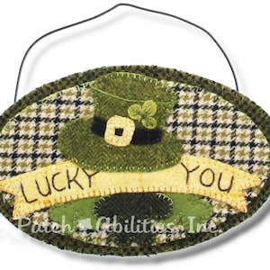 May include: A green felt oval ornament with a green leprechaun hat, a yellow banner with the text "LUCKY YOU", and a green plaid background.