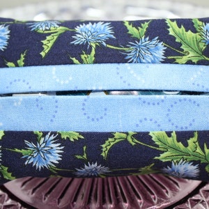 May include: A blue and white fabric tissue box cover with a floral pattern. The cover has two blue stripes and a white background with blue dots.