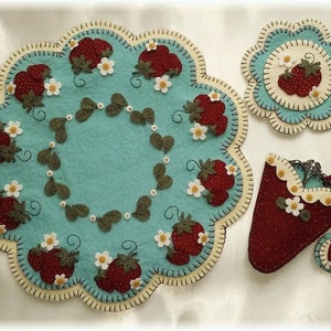 May include: A set of three felt crafts featuring a strawberry theme. The set includes a round placemat with a blue background and a border of white and yellow felt, a smaller round coaster with a similar design, and a strawberry-shaped needle case with a red felt body and a white felt trim.