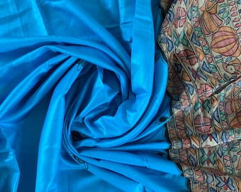 May include: A blue silk saree with a colorful, intricate border design. The saree is draped and folded, showcasing the fabric's texture and the vibrant border pattern.
