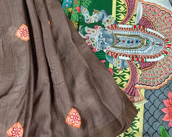 May include: A brown cotton saree with a floral design. The saree is draped over a colorful fabric with a painted scene of a deity with a cow and a tree.