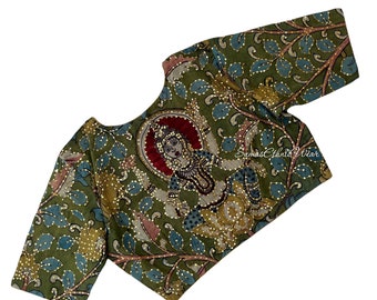May include: A green, blue and gold embroidered blouse with a floral pattern and a depiction of a deity. The blouse has a round neckline and three-quarter sleeves. The text "Sumas Ethnic Wear" is visible in the bottom right corner.