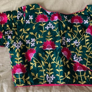 May include: A green blouse with long sleeves and a pink trim. The blouse is embroidered with pink and white flowers and green leaves.