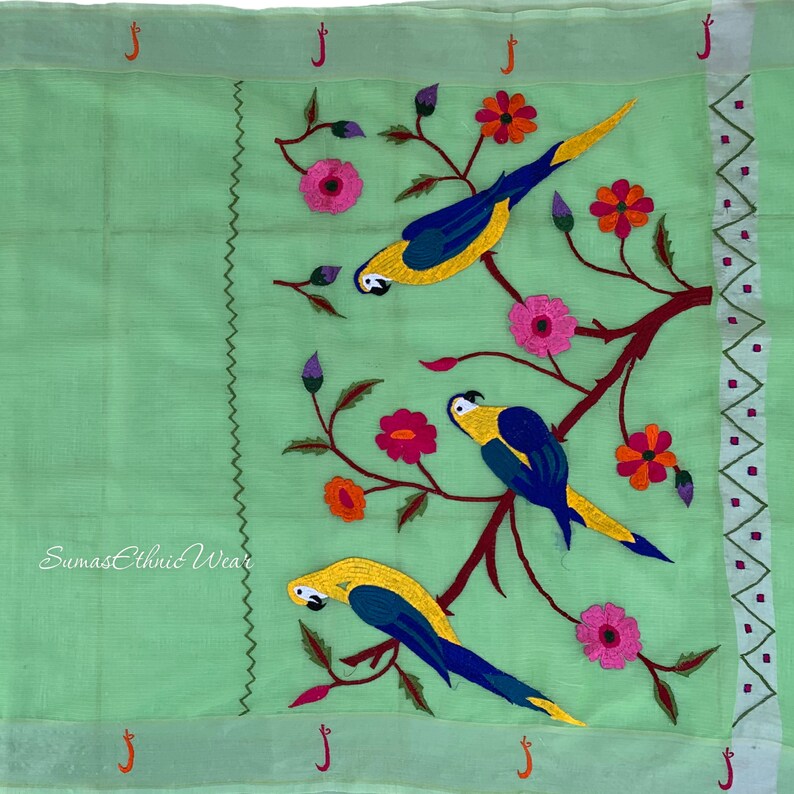 May include: A green fabric with embroidered parrots and flowers. The parrots are yellow, blue, and green. The flowers are pink, red, and orange. The fabric has a white border with a repeating pattern of small squares.