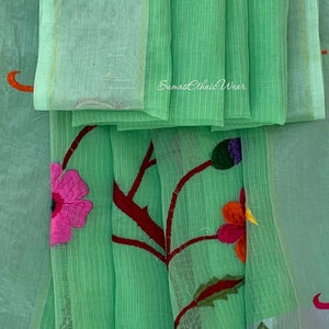 May include: A light green sheer fabric with embroidered flowers in pink, red, orange, yellow, and purple. The fabric has a subtle striped pattern and the text "Sumas Ethnic Wear" is visible in the top right corner.