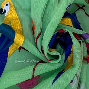 May include: Green fabric with embroidered parrots and flowers. The fabric has a subtle texture and is sheer. The text "Sumas Ethnic Wear" is visible in the bottom left corner.