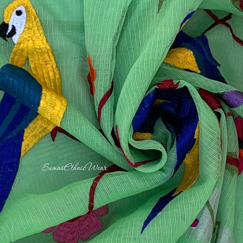 May include: Green fabric with embroidered parrots and flowers. The fabric has a subtle texture and is sheer. The text "Sumas Ethnic Wear" is visible in the bottom left corner.