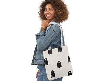 May include: A white tote bag with a black ghost pattern. The bag has white straps and is being held by a person wearing a denim jacket and jeans.