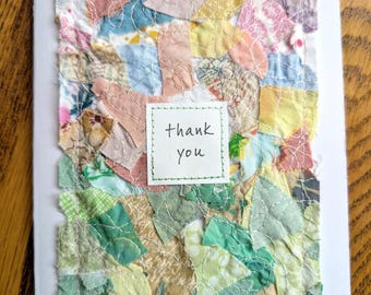 Puede incluir: Una tarjeta de felicitación blanca con un collage de retazos de tela multicolores en tonos verdes, rosas, amarillos y azules, asegurados con costuras decorativas blancas. Una etiqueta cuadrada central muestra el texto thank you en negro.