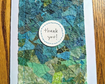 Puede incluir: Una tarjeta de felicitación rectangular que presenta un collage de varios retazos de tela azul y verde cosidos con hilo azul. Una etiqueta circular de papel blanco está centrada sobre la tela, mostrando el texto escrito a mano thank you!