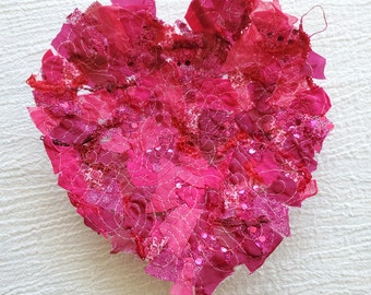 Puede incluir: Una obra de arte de tela en forma de corazón rosa hecha de muchos pequeños trozos de tela cosidos juntos. La tela es una mezcla de rosa, rojo y blanco, y el corazón está decorado con lentejuelas.