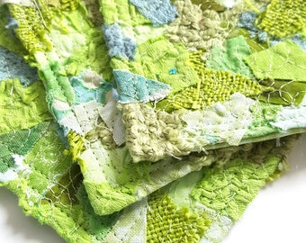 Puede incluir: Un juego de cuatro posavasos de tela verde y blanca con un diseño de patchwork. Los posavasos están hechos de diferentes telas y texturas, y están cosidos juntos con hilo blanco.