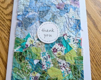 Puede incluir: Tarjeta de felicitación hecha a mano con un diseño de patchwork. La tarjeta presenta una superficie texturizada con piezas de tela azul, verde y floral cosidas juntas. Una etiqueta circular blanca con las palabras "gracias" está centrada en la tarjeta.