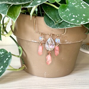May include: A copper-coloured plant pot hanger with a clear crystal teardrop and pink crystal accents. The hanger also features iridescent beads and pink crystal points. The plant pot is filled with a green plant with silver-speckled leaves.