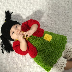 May include: A baby wearing a handmade crochet costume. The costume includes a black hat with long pigtails, a red jacket, a green dress with a yellow square, and white shorts. The baby is lying on a white, textured surface.