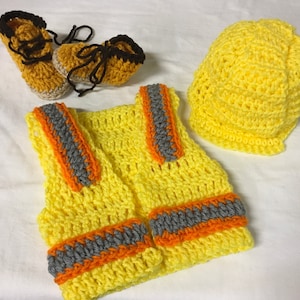 May include: A crocheted construction worker costume set for a baby. The set includes a yellow vest with orange and gray stripes, a yellow hard hat, and a pair of brown and black booties.