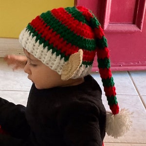 May include: A child wearing a red, green, and white striped elf hat with a long tail and elf ears. The hat is crocheted and has a white pom-pom on the end of the tail.