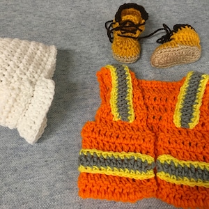 May include: A crocheted construction worker costume set for a baby. The set includes a white hat, an orange vest with yellow and gray reflective stripes, and a pair of brown and yellow booties.