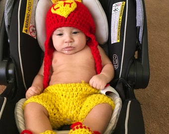 May include: A baby wearing a red and yellow crocheted superhero costume with a heart-shaped symbol on the chest. The costume includes a hat, shorts, and booties. The baby is sitting in a car seat.