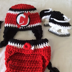 May include: A crocheted baby hat, diaper cover, and booties set in red, black, and white with a New Jersey Devils logo.