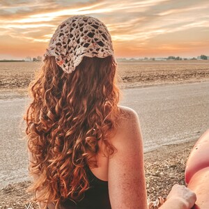 May include: A woman with long, curly red hair wearing a crocheted hat stands in a field with a sunset in the background.