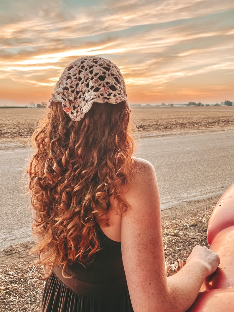 May include: A woman with long, curly red hair wearing a crocheted hat stands in a field with a sunset in the background.