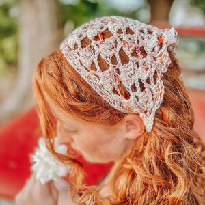 May include: A woman with long, curly red hair is wearing a white crocheted headband with a colorful pattern. She is holding a white flower and smelling it.