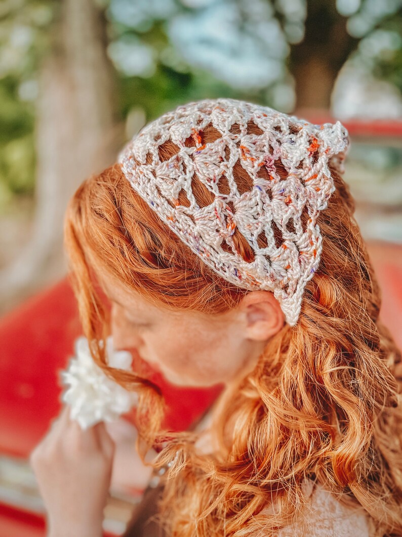 May include: A woman with long, curly red hair is wearing a white crocheted headband with a colorful pattern. She is holding a white flower and smelling it.